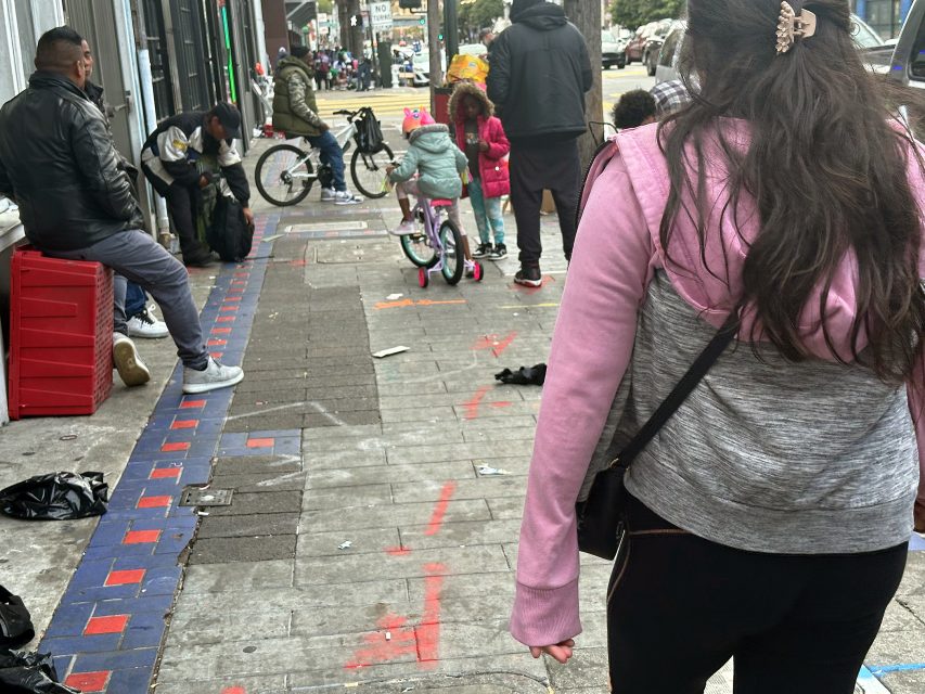 People, including children with bicycles, gather on a city sidewalk with scattered belongings and some sitting near a building. The street and cars are visible in the background.