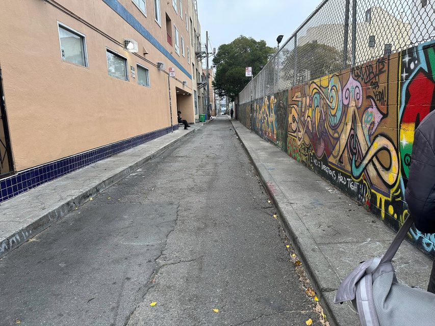 A narrow urban alleyway bordered by a building on the left and a colorful graffiti-covered wall on the right, with overcast sky and scattered trash bins in the distance.