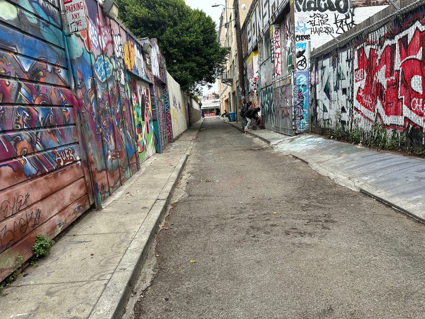 Narrow urban alleyway with walls and gates covered in colorful graffiti; a couple of people are visible in the distance.