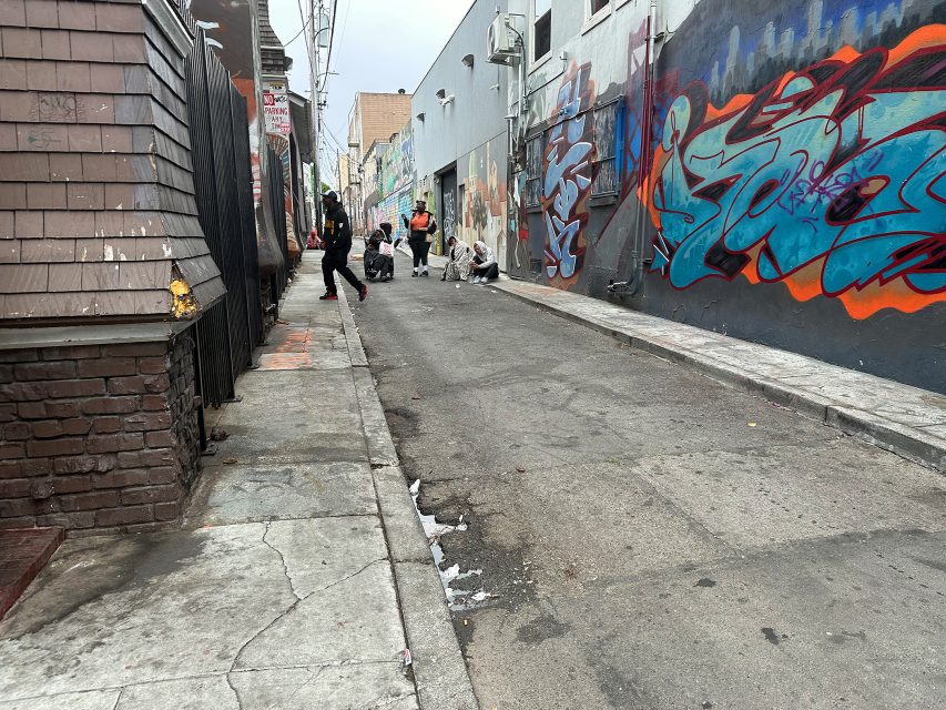 A group of people are gathered on a graffiti-covered alleyway, with some kneeling on the ground and others standing nearby. The pavement is cracked and littered with debris.