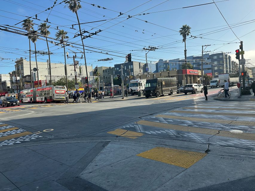 Urban intersection with buses, cars, and pedestrians crossing; overhead wires and palm trees visible, with buildings in the background under a clear sky.