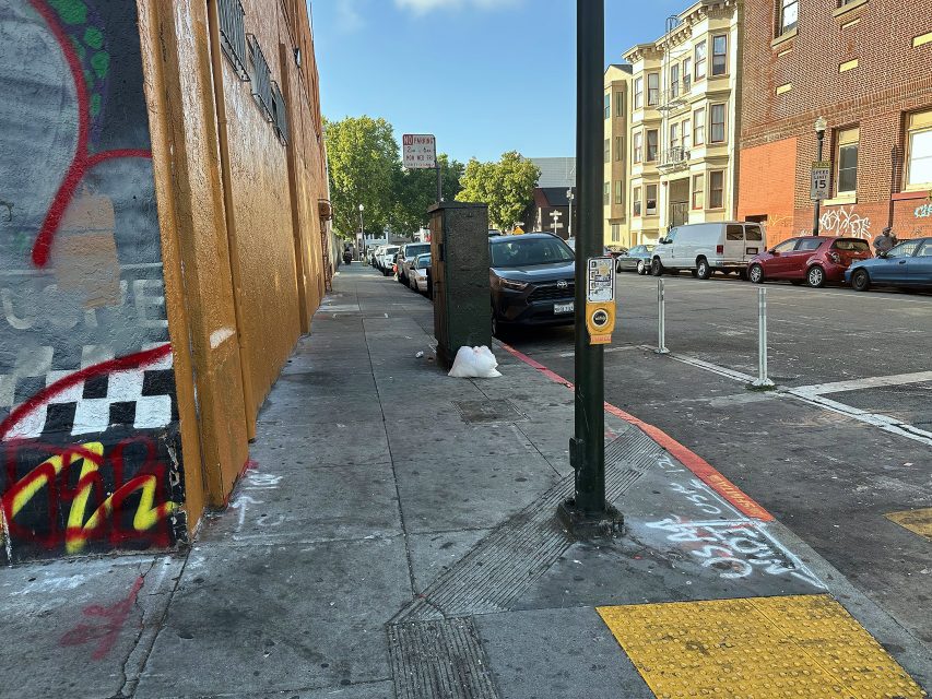A city sidewalk with graffiti, a crosswalk button, parked cars, a bag of trash by the curb, and apartment buildings in the background.