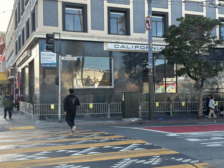 Street corner with a building displaying a "For Lease" sign, metal barricades along the sidewalk, and pedestrians crossing at a crosswalk with yellow markings.