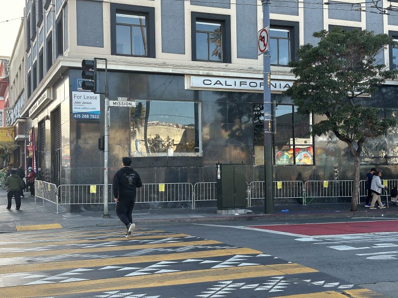 Street corner with a building displaying a "For Lease" sign, metal barricades along the sidewalk, and pedestrians crossing at a crosswalk with yellow markings.