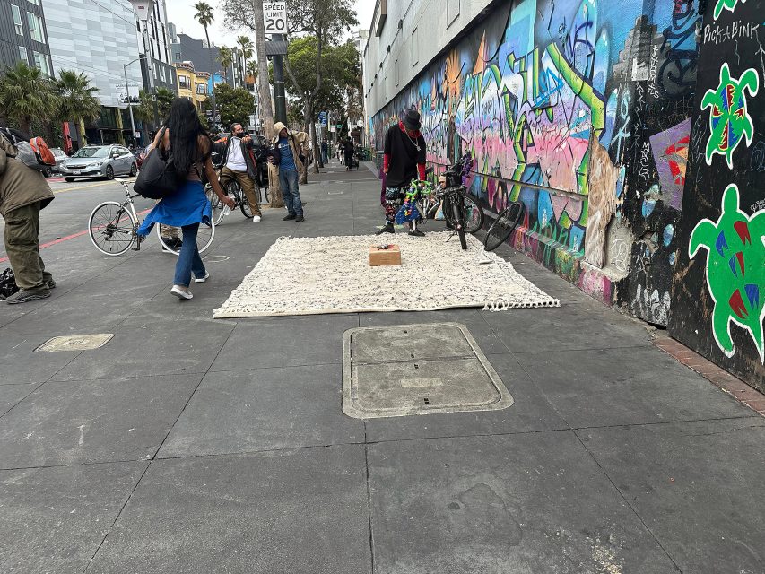 A person stands beside a bicycle near a wall with colorful graffiti; a large white rug with a box on it is spread on the sidewalk.