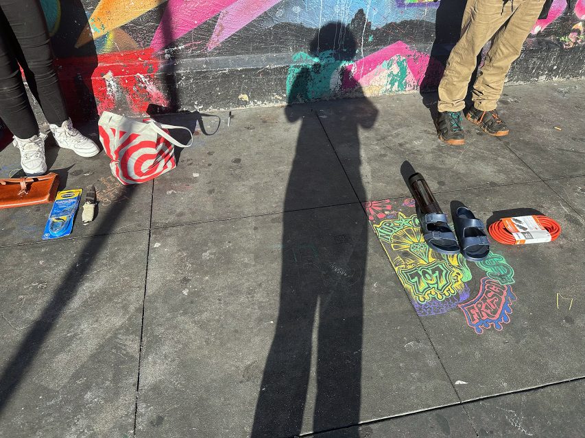 Two people stand near a colorful chalk drawing on a sidewalk with various items on the ground; a shadow of a photographer is cast in the center.