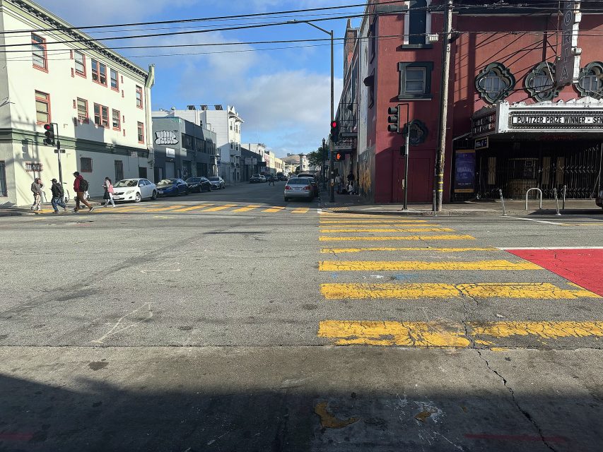 A city street intersection with yellow crosswalk lines, a few cars, pedestrians crossing, and buildings on both sides under a partly cloudy sky.