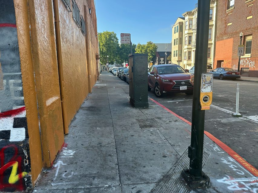 City sidewalk with graffiti on the curb, a pedestrian crossing button on a pole, parked cars along the street, and buildings in the background under clear skies.