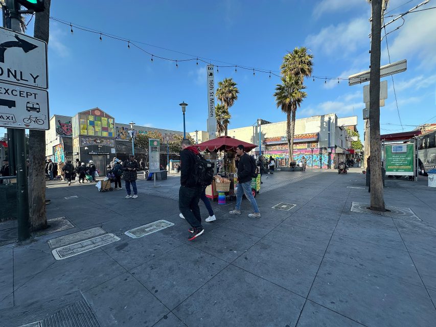 Street scene with people walking past a vendor cart under a red umbrella; palm trees, murals, and various buildings in the background on a sunny day.