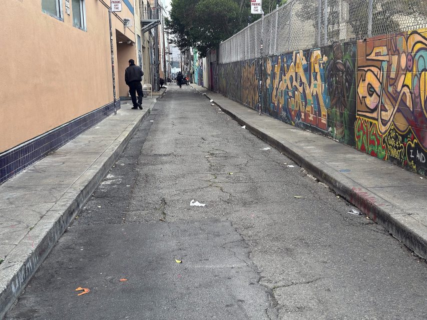 A narrow urban alley with graffiti-covered walls, scattered litter, and a few people walking in the distance.
