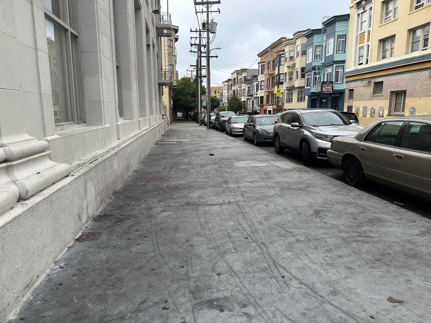A city sidewalk lined with parked cars and multi-story buildings on a cloudy day. The street is mostly empty, with no people visible.