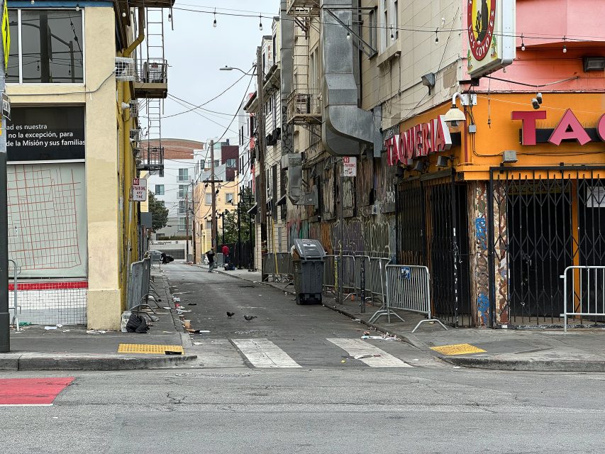 A narrow, empty urban alley with scattered trash, closed storefronts, metal barricades, and a few pigeons on the ground during daylight.