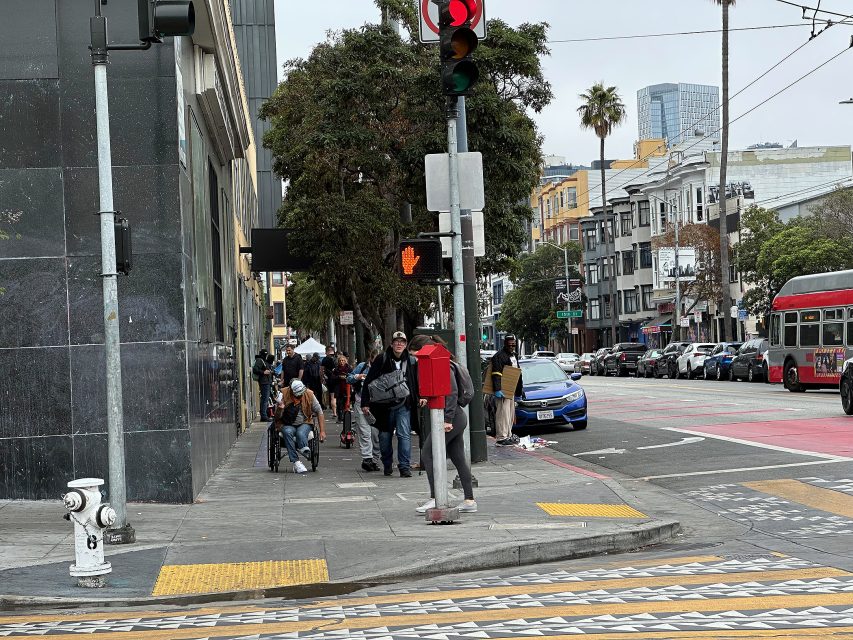 A group of people stand and sit on a city sidewalk near a street corner; cars and a bus are visible along the road on a cloudy day.