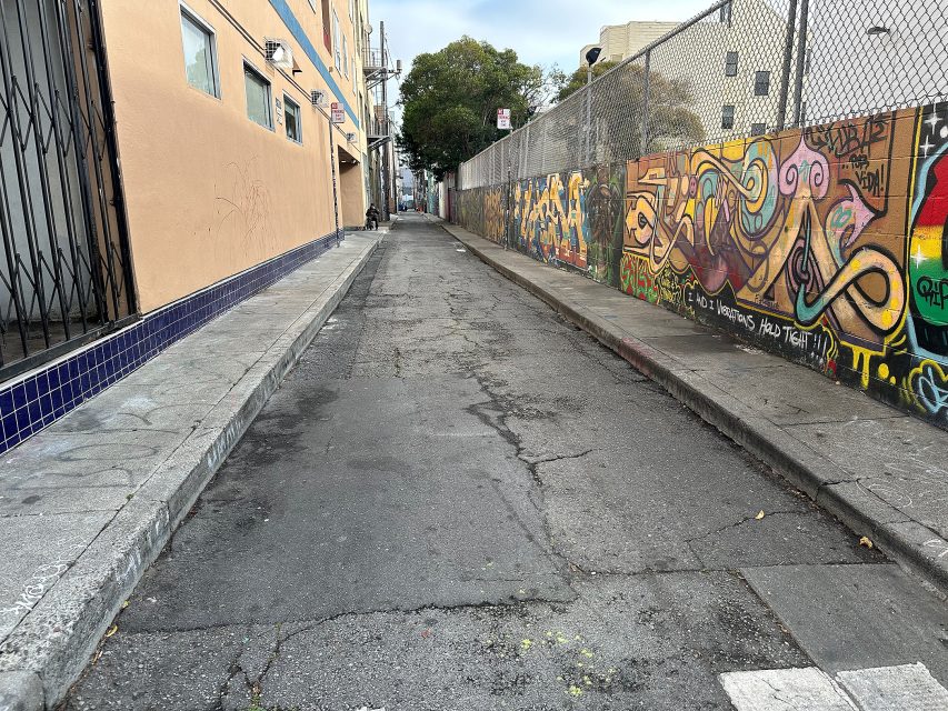 A narrow urban alleyway with cracked pavement, a beige building on the left, and a chain-link fence with colorful graffiti on the right.