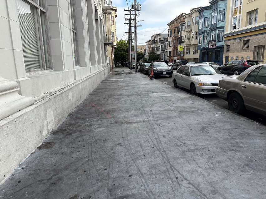 A city sidewalk with parked cars along the street, multi-story buildings on both sides, and power lines overhead on a cloudy day.