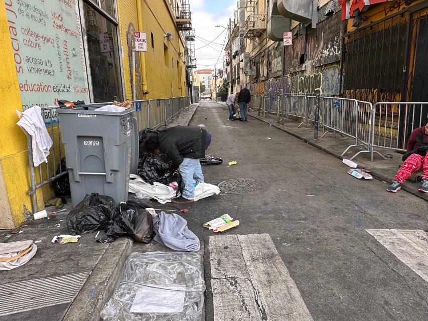 A person searches through a trash bin in an alley with scattered debris, while other people stand and sit nearby; buildings and graffiti line the street.