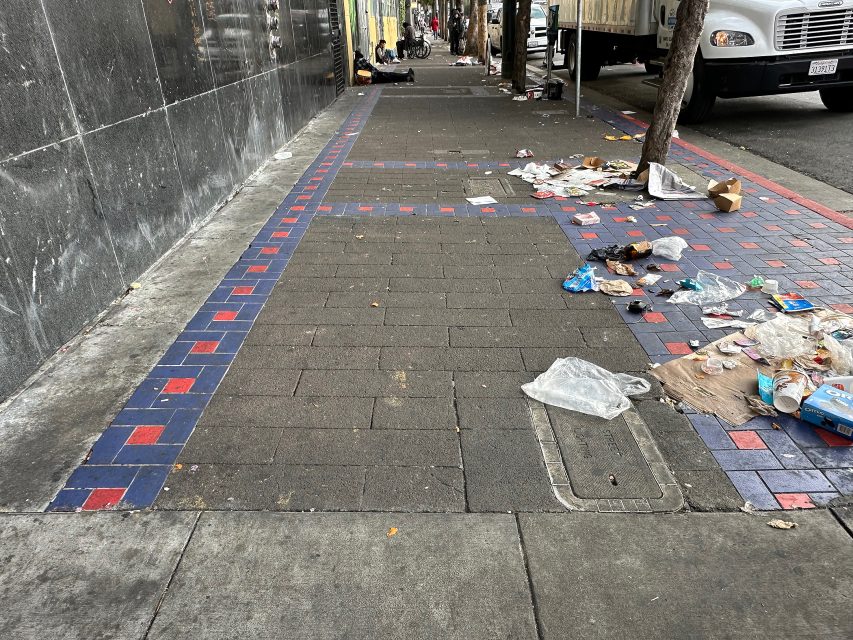 A city sidewalk with scattered litter and debris, bordered by a building wall and street trees. A truck and pedestrians are visible in the background.