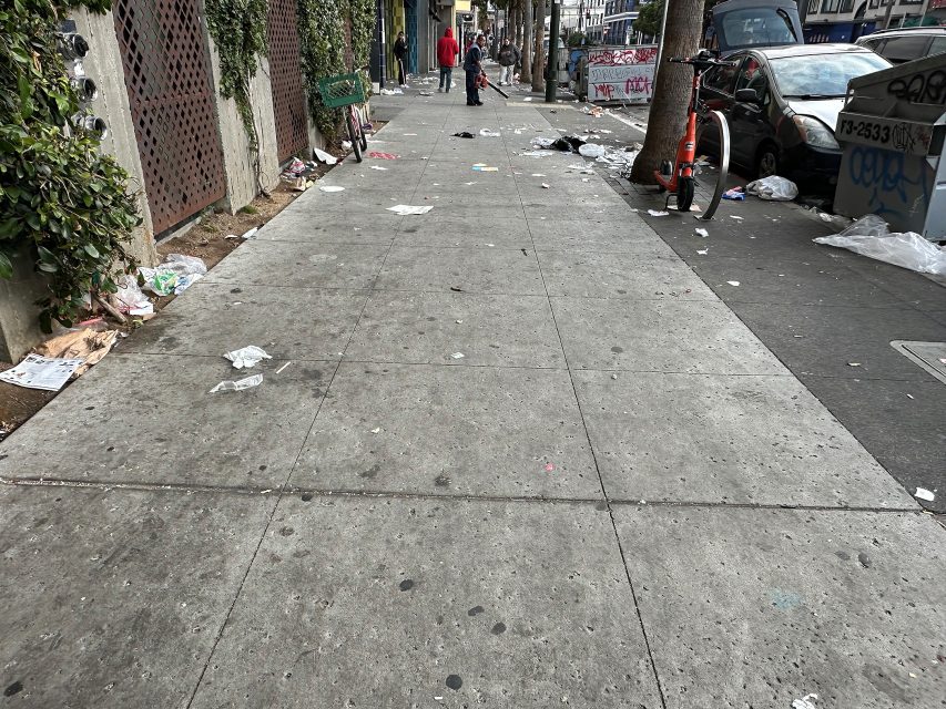 A city sidewalk littered with trash, with a few people in the distance, a parked bicycle, and cars along the street.