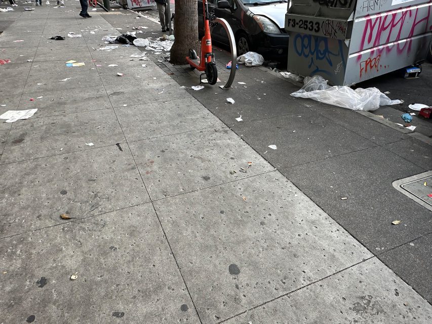 A city sidewalk with scattered litter, a parked electric scooter, graffitied walls, and a covered car.