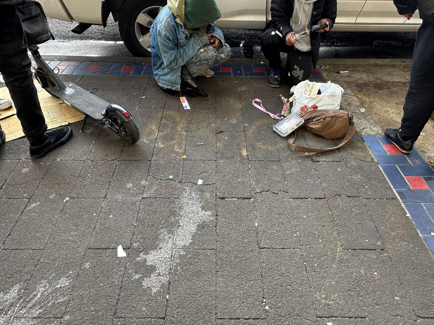 Two people sit on a city sidewalk near a parked scooter, surrounded by scattered belongings, as others stand nearby.
