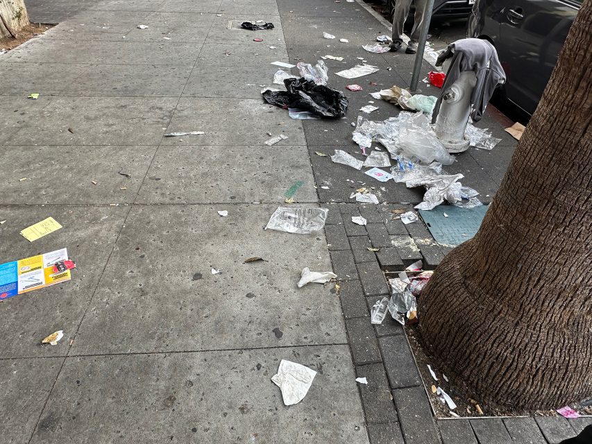 Trash and litter are scattered across a city sidewalk and street near a parked car and a tree.