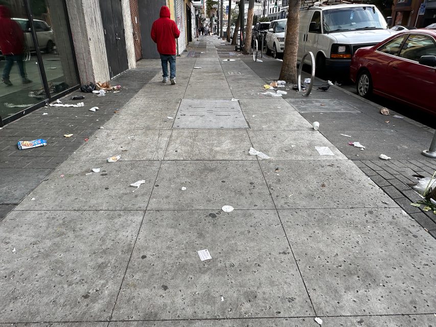 A person in a red hoodie walks down a city sidewalk littered with trash, with parked cars and storefronts along the street.
