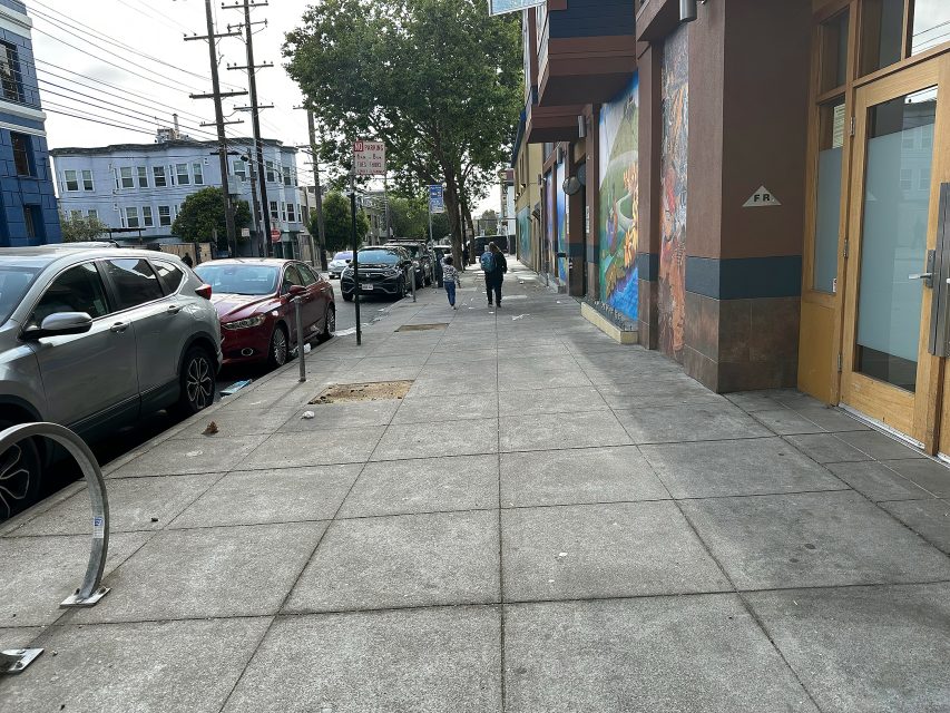 A city sidewalk with parked cars, a mural on one building, and two people walking in the distance. Trees and utility poles line the street.