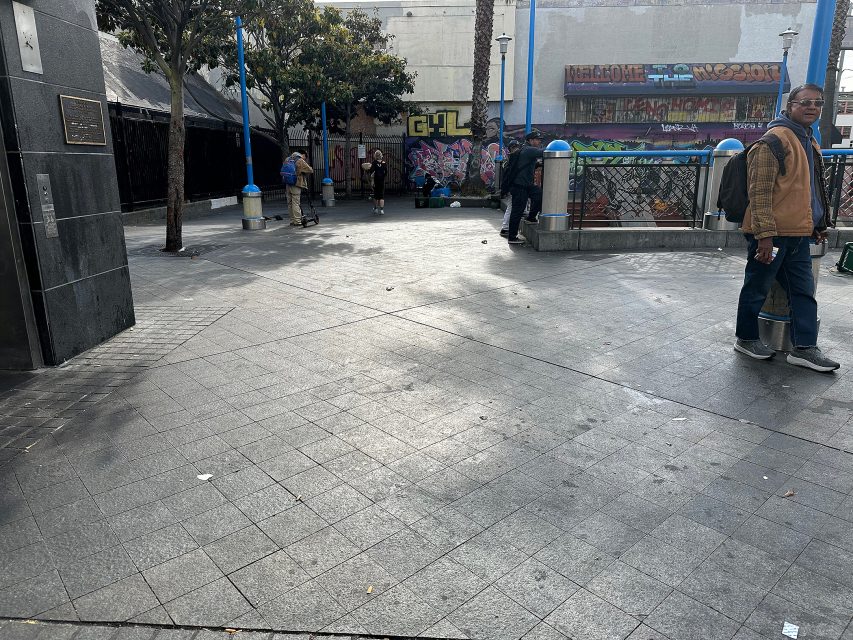 Outdoor urban plaza with several people standing and walking; graffiti-covered walls and a stairway entrance are visible in the background.