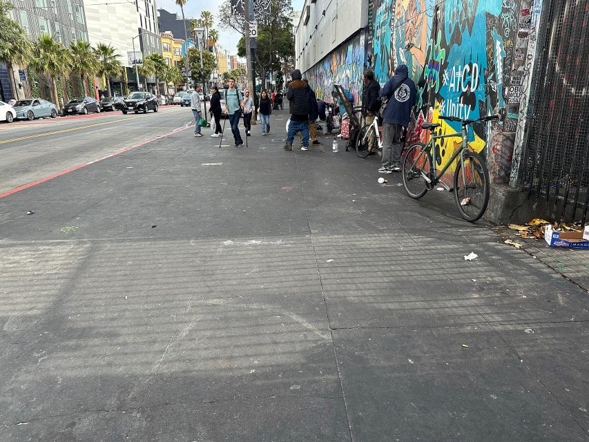 People walking and standing near a colorful graffiti-covered wall on a city sidewalk; bicycles are parked against the wall and cars are parked along the street.