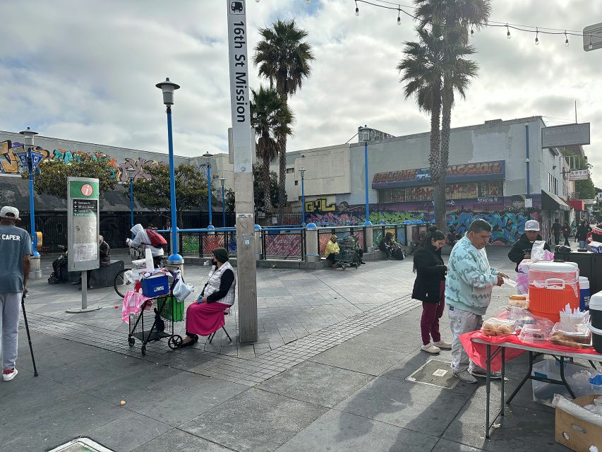 People gather near the 16th St Mission BART station; some are seated, others stand by food and drink tables, with graffiti and palm trees in the background on a cloudy day.