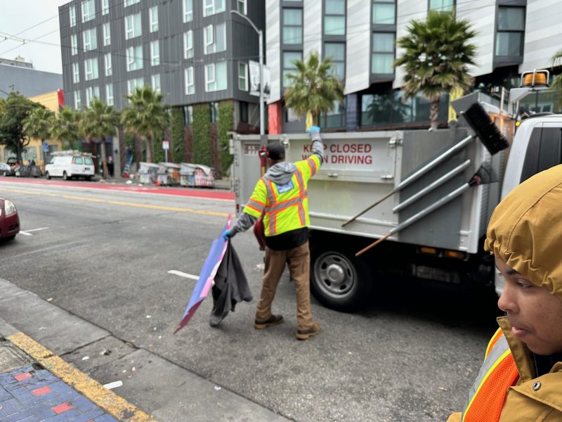 A worker in a reflective jacket stands near a utility truck on a city street, holding a folded sign and jacket. Another person wearing a hood is visible in the foreground.