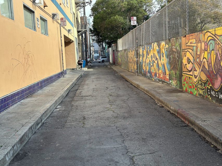 Narrow urban alleyway with graffiti-covered wall on the right and a yellow building on the left. Sidewalks border the cracked pavement; a person is visible in the distance.
