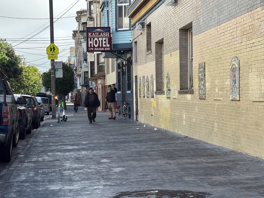 A city sidewalk with pedestrians, parked cars, scooters, and a sign for Kailash Hotel at 179 Julian Ave; yellow crosswalk sign and wall murals are visible.