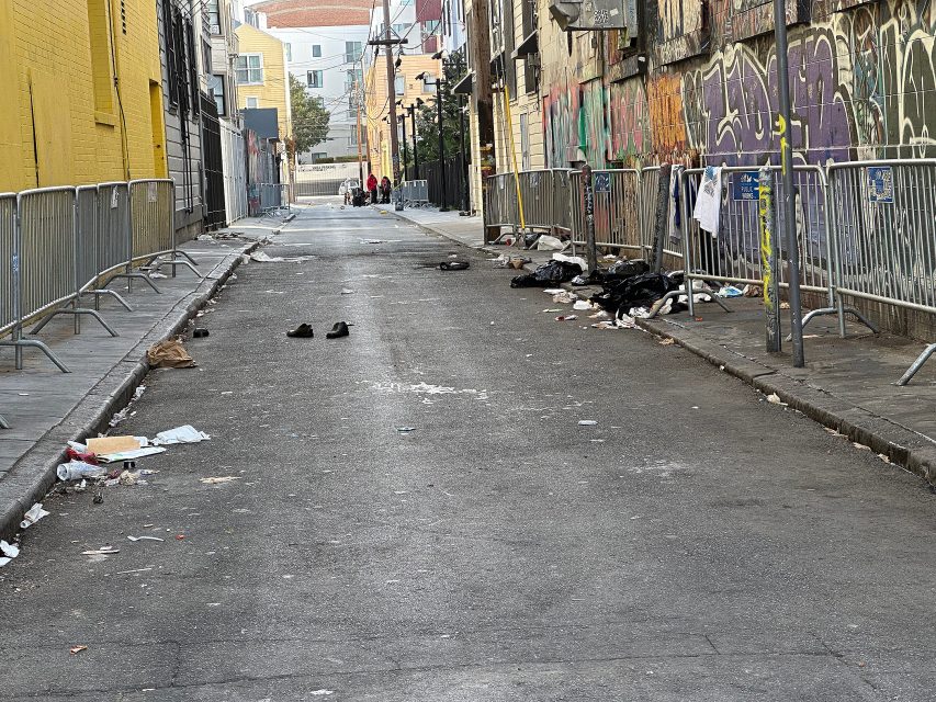 A deserted urban alleyway with scattered trash, fencing on both sides, graffiti on one wall, and a few people visible in the distance.