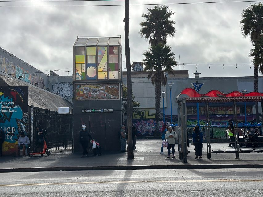 A city street scene shows people waiting near a bus stop, with colorful graffiti on walls and a modern glass structure above a building entrance. Palm trees and cloudy sky are visible.