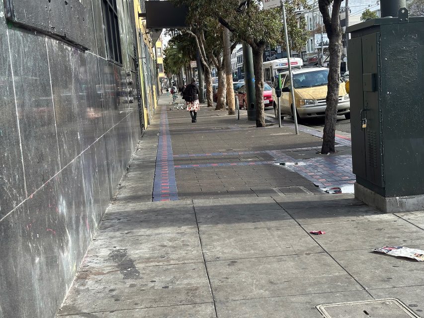 A city sidewalk with scattered litter, a tree-lined street, parked cars, a utility box, and a person walking away in the distance.