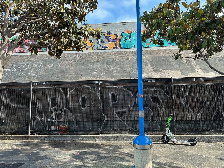 A sidewalk with a parked electric scooter, trees, and a building covered in graffiti and tags on the wall and roof.