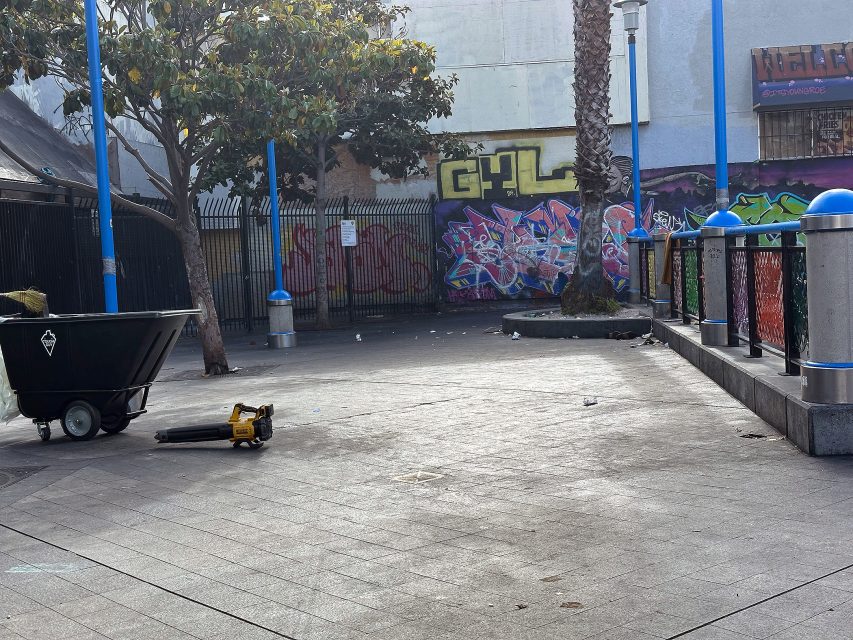 A mostly empty urban plaza with graffiti on the walls, a large trash bin, a leaf blower on the ground, and a few trees and blue lampposts.