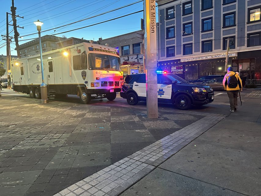 Two police vehicles, including a large command truck and an SUV with lights on, are parked near the 16th St Mission BART station. A person with a backpack walks nearby.
