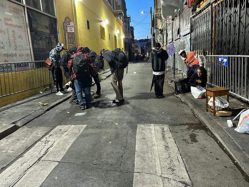 A group of people gather and sit on the sidewalk in an urban alley lined with graffiti and litter in the evening.