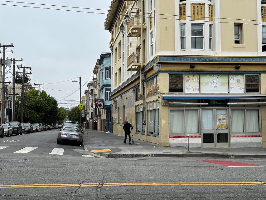 A person stands on a sidewalk near the corner of a street lined with parked cars and buildings under an overcast sky.