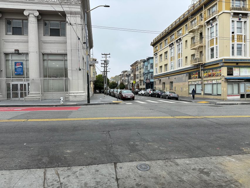 A city street intersection with parked cars, multi-story buildings, and a crosswalk on a cloudy day.