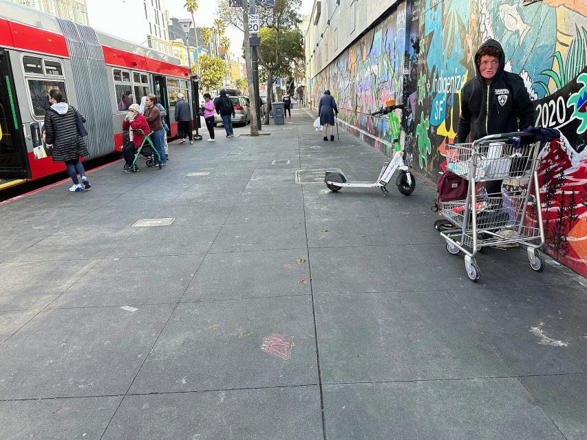 A man with a shopping cart stands by a colorful graffiti wall; people board a bus on the left, and electric scooters are parked on the sidewalk.