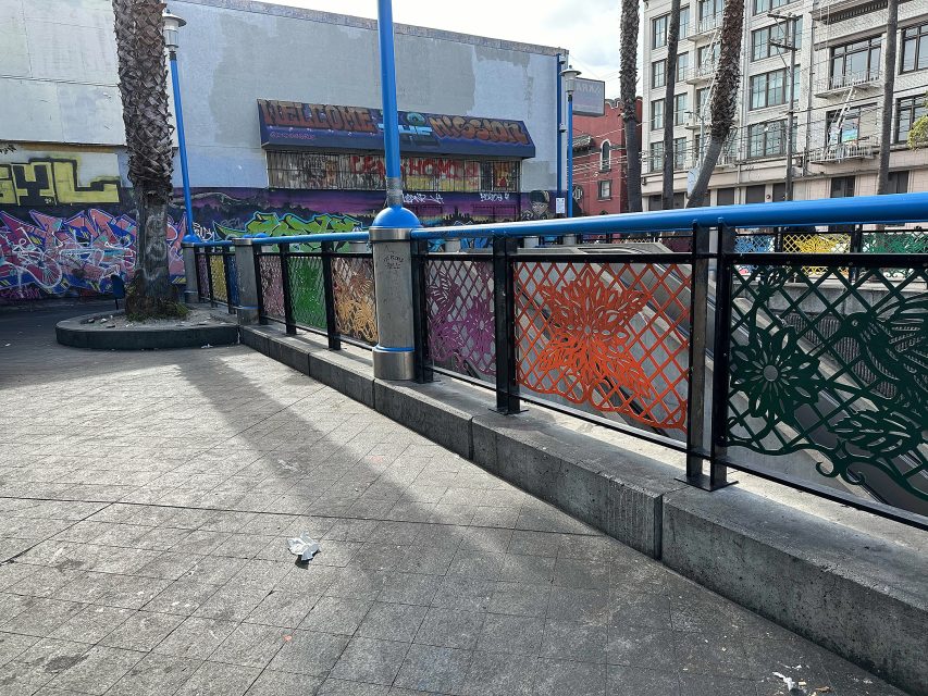 Urban park scene with decorative metal fence panels, street art on walls, palm trees, lamp posts, and multi-story buildings in the background.