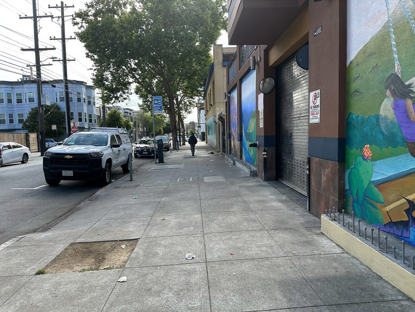 A city sidewalk with parked cars, a few pedestrians, and colorful murals on the building walls. Trees line the street and utility poles are visible.