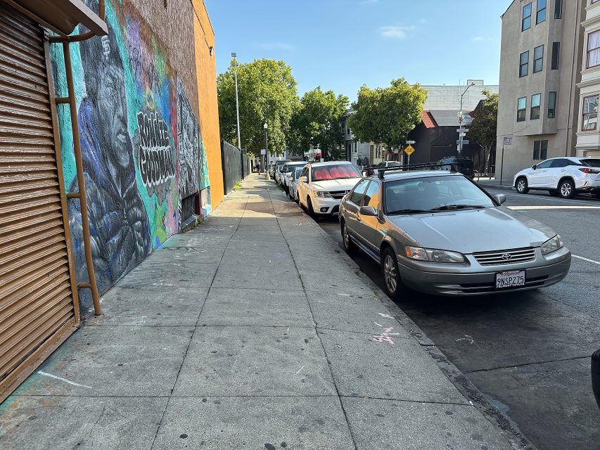 A city sidewalk with parked cars, a mural on the left wall, and buildings lining both sides of the street on a clear day.