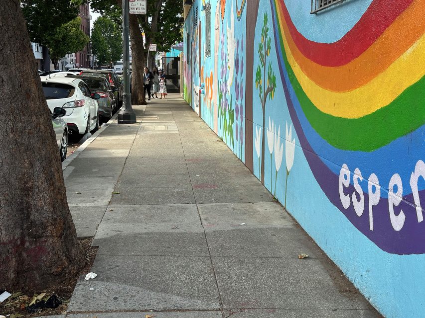 A sidewalk lined with parked cars runs beside a mural-painted wall featuring a rainbow and white flowers; a tree is in the foreground.