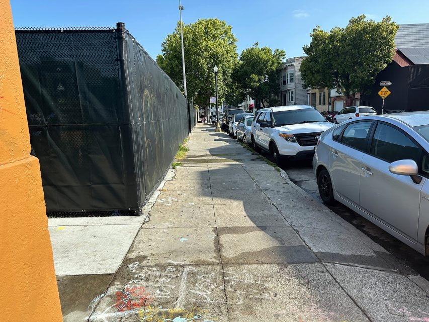 A city sidewalk with parked cars on the right, a black fence on the left, some chalk drawings on the pavement, and trees and buildings in the background under a blue sky.