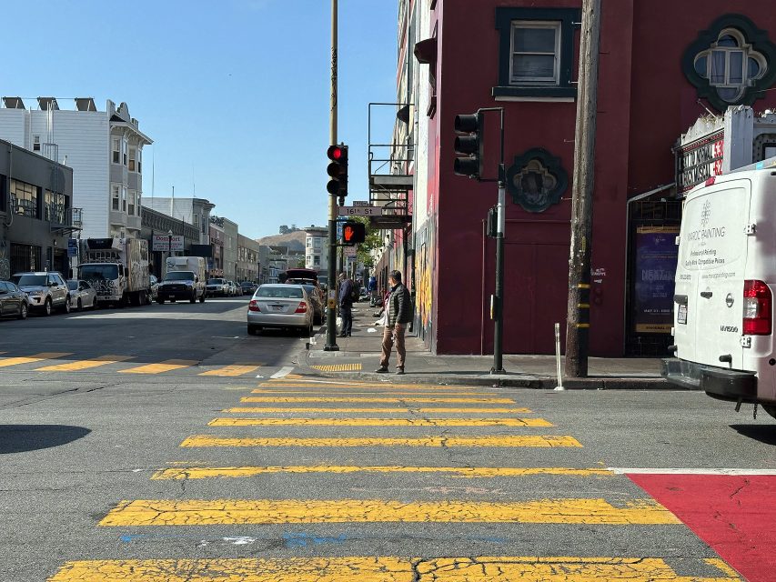 A city intersection with yellow crosswalk lines, a red traffic light, pedestrians waiting to cross, and vehicles parked along the street on a sunny day.