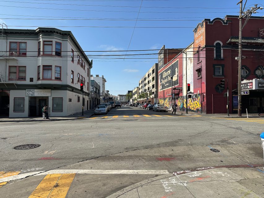 A city street intersection with crosswalks, buildings on both sides, some parked cars, murals, and a few pedestrians under a clear blue sky.